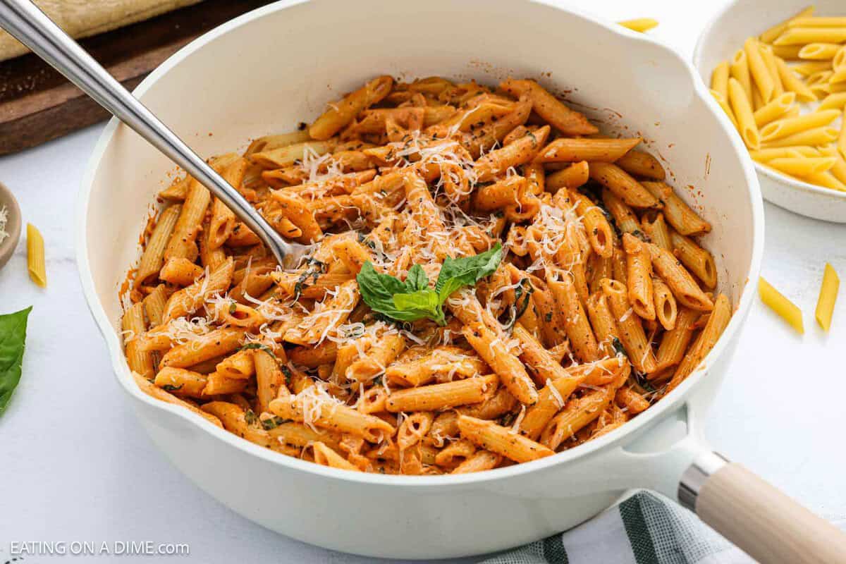 A white skillet filled with Penne alla Vodka in a creamy tomato sauce, topped with grated cheese and fresh basil, with a spoon inside.