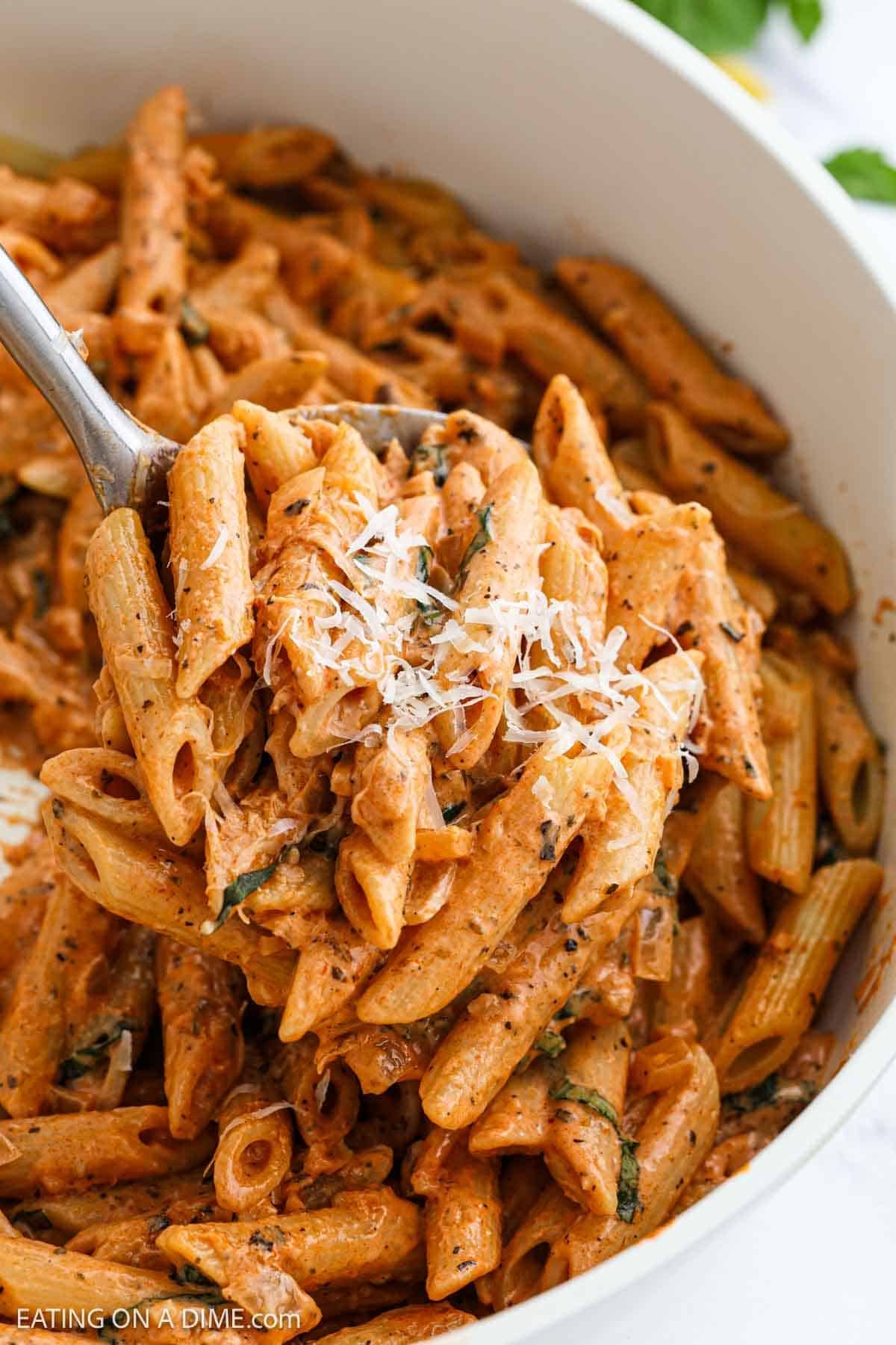 A close-up of creamy pasta dish, garnished with grated cheese and fresh herbs. A serving spoon is lifting a portion of the pasta from the dish.