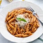 A white bowl filled with Penne alla Vodka in a creamy vodka sauce, topped with grated cheese and a sprig of basil, with a fork resting on the bowl’s edge.