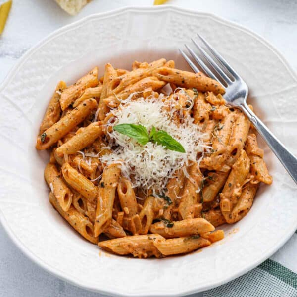 A white bowl filled with Penne alla Vodka in a creamy vodka sauce, topped with grated cheese and a sprig of basil, with a fork resting on the bowlโs edge.