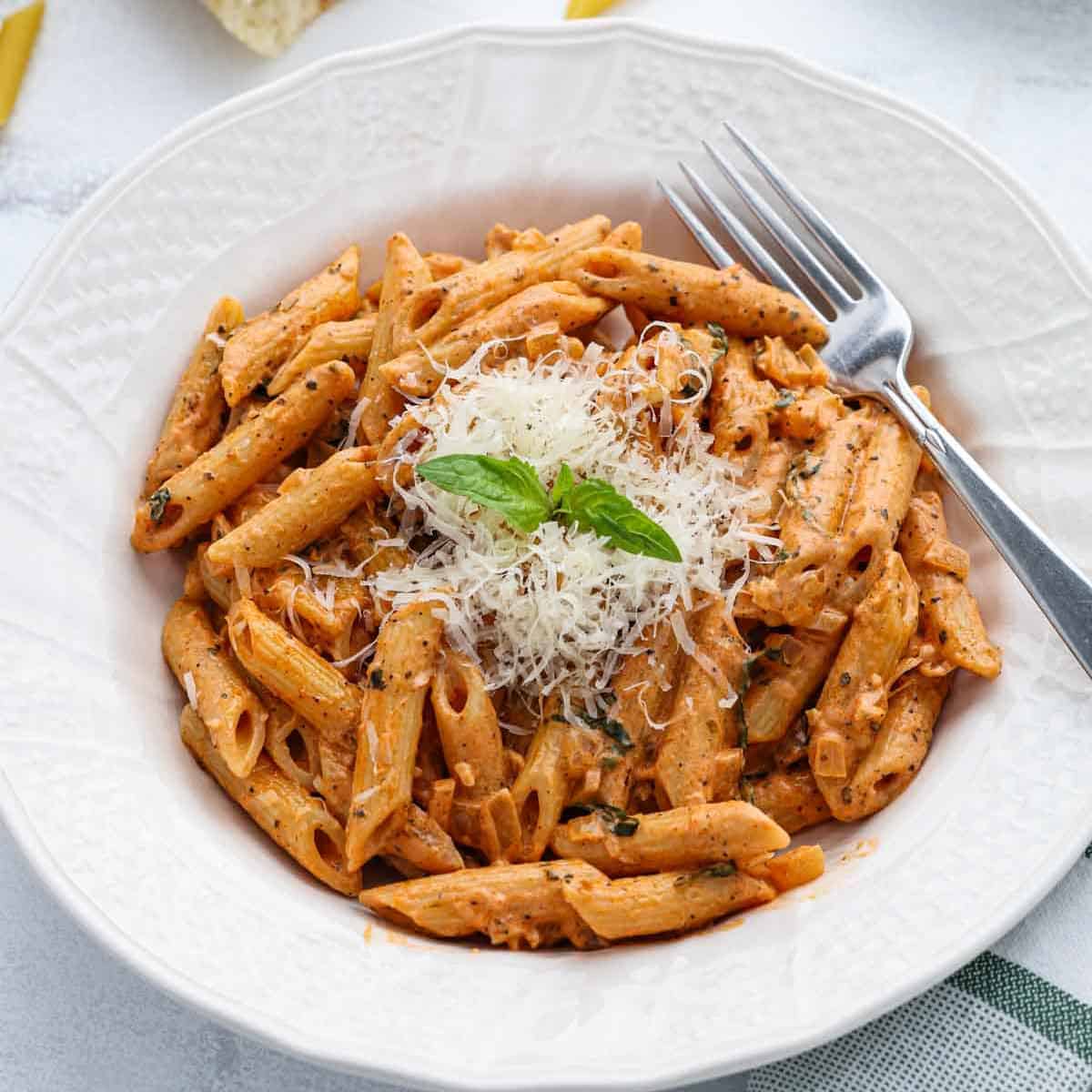 A white bowl filled with Penne alla Vodka in a creamy vodka sauce, topped with grated cheese and a sprig of basil, with a fork resting on the bowl’s edge.
