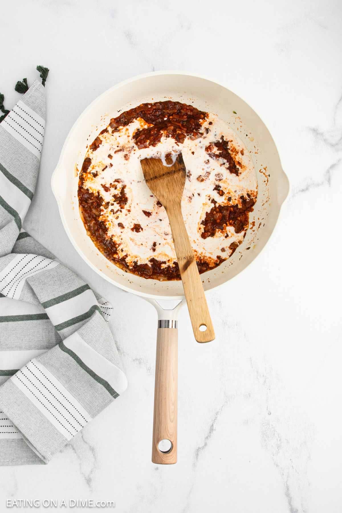 A white skillet with cooked tomato paste and seasoning mixed together with the heavy cream with a wooden spoon.