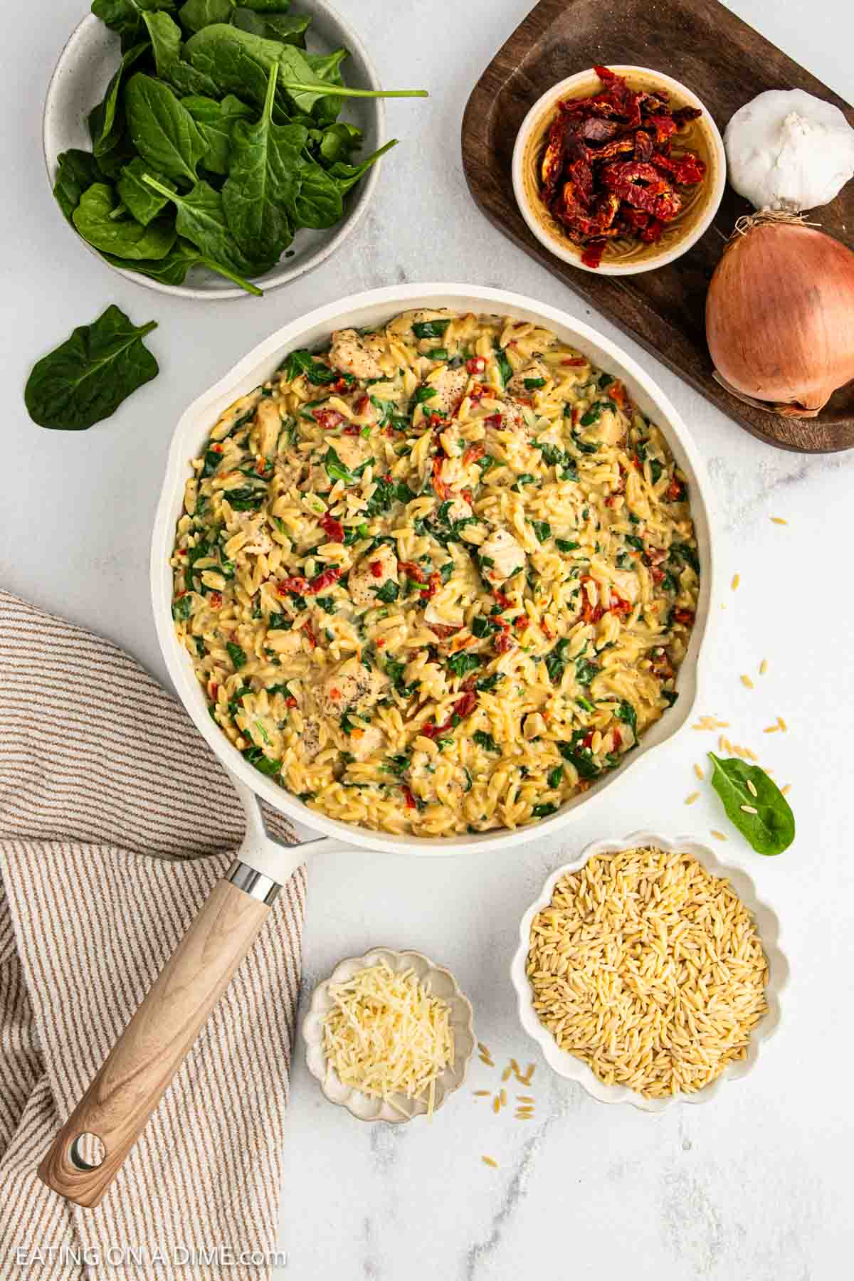 A Tuscan chicken orzo skillet filled with creamy orzo pasta, spinach, chicken, and sun-dried tomatoes sits on a table. Surrounding it are fresh spinach, an onion, a bowl of sun-dried tomatoes, uncooked orzo, and grated cheese—perfect for your next recipe.