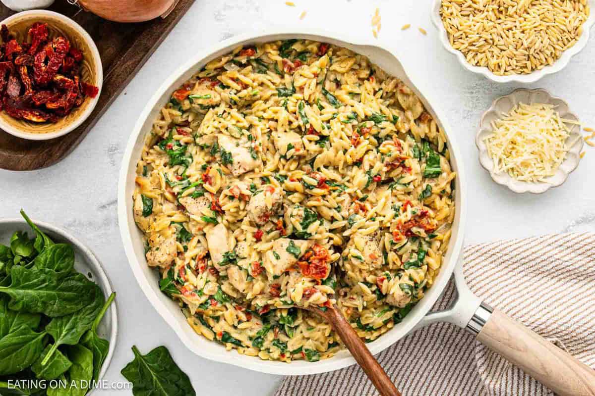 A delicious orzo skillet filled with creamy orzo pasta, Tuscan chicken, spinach, and sun-dried tomatoes, with a wooden spoon inside. Nearby are bowls of dry orzo, grated cheese, sun-dried tomatoes, and fresh spinach leaves.
