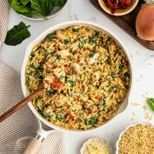 A skillet filled with creamy orzo pasta, Tuscan chicken, spinach, and sun-dried tomatoes, with a wooden spoon resting inside. Ingredients like spinach leaves, an onion, orzo, and cheese are arranged nearby for this flavorful recipe.