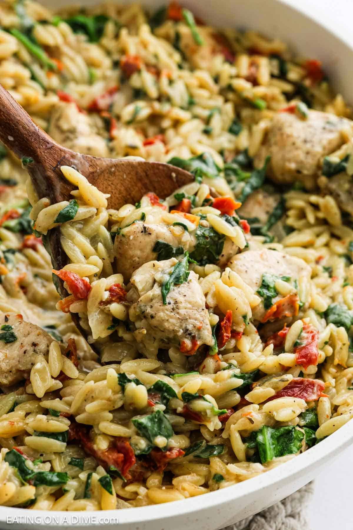 A wooden spoon scoops creamy Tuscan chicken orzo skillet, with tender chicken, spinach, and sun-dried tomatoes from a bubbling pan—a comforting recipe for any night.