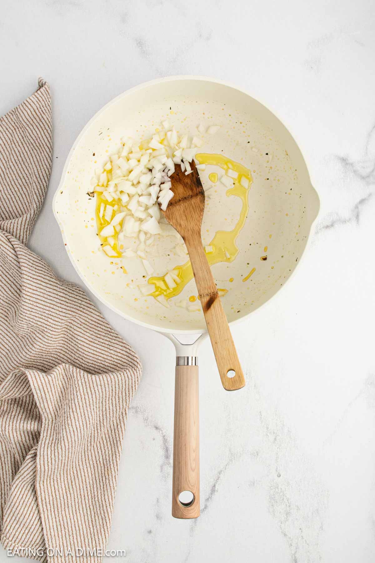 A white skillet for your Tuscan chicken recipe holds chopped onions and olive oil, stirred with a wooden spatula on a white marble surface beside a beige striped kitchen towel.