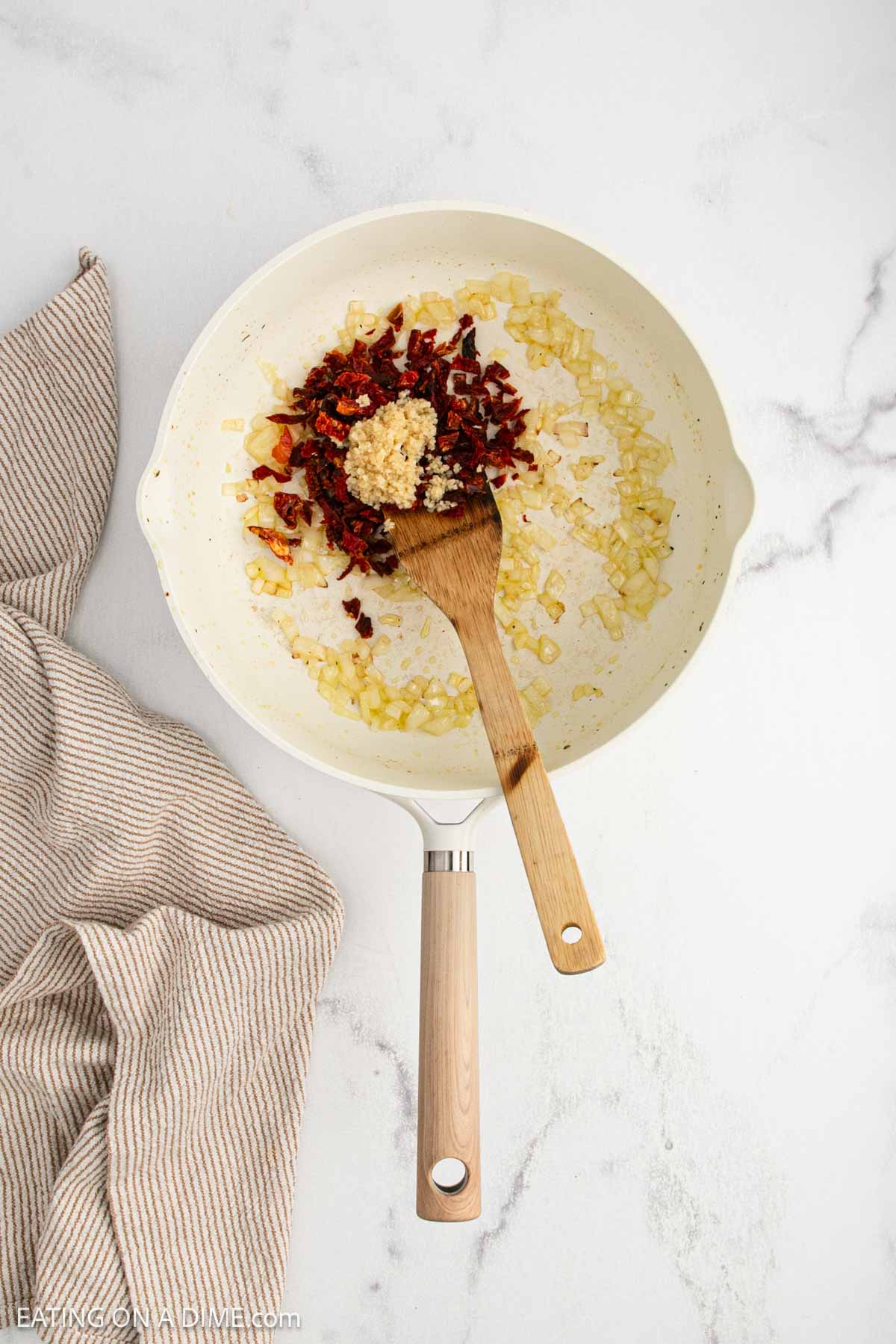 A white skillet on a marble surface holds ingredients for a Tuscan chicken orzo skillet—sautéed onions, sun-dried tomatoes, and minced garlic—stirred with a wooden spoon. A beige striped kitchen towel rests nearby in this inviting recipe scene.