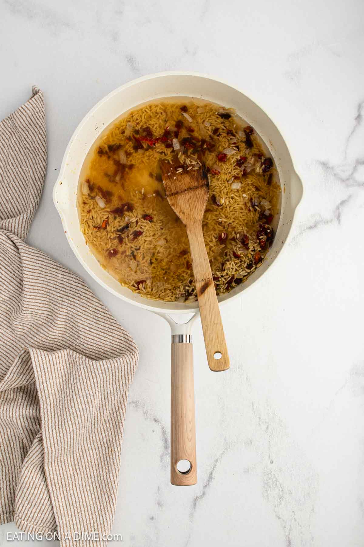 A white skillet on a marble surface holds rice, broth, and spices being stirred with a wooden spatula—a cozy scene reminiscent of a classic Tuscan chicken recipe. A beige-striped kitchen towel lies next to the skillet.