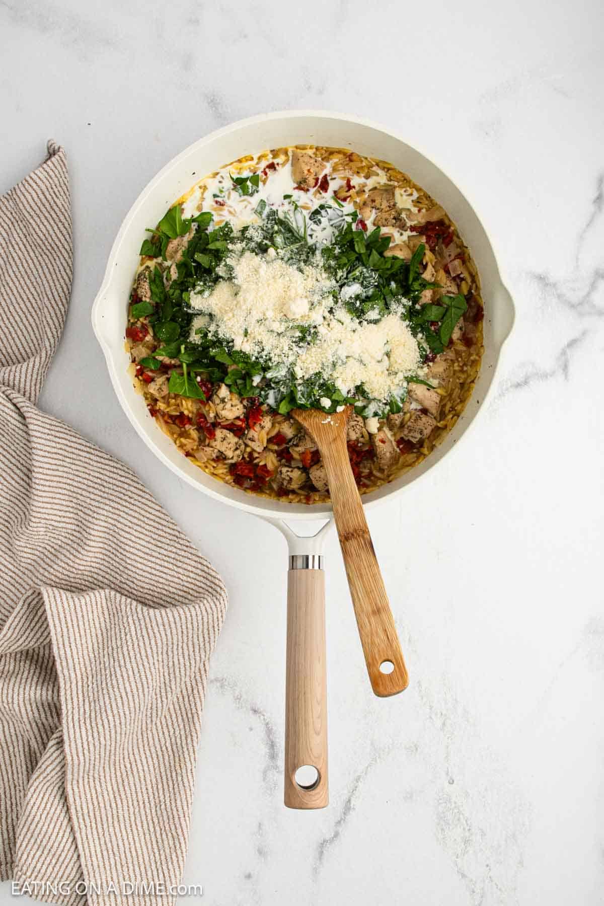 A creamy orzo skillet recipe with fresh spinach and grated cheese, served in a skillet with a wooden spoon. The dish rests on a marble surface beside a beige striped towel, perfect for your next cozy dinner.