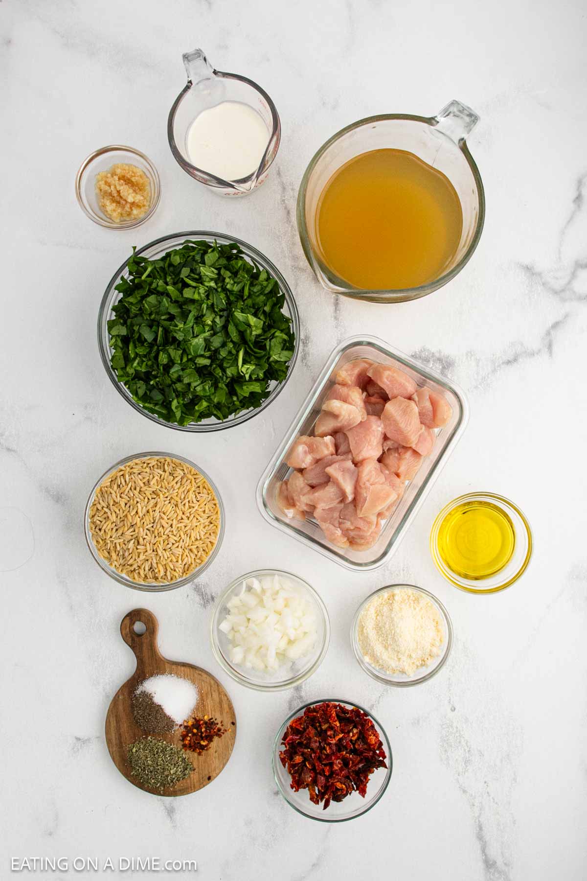 Various ingredients are arranged on a white marble surface for a Tuscan Chicken Orzo Skillet, including chopped spinach, cubed raw chicken, orzo pasta, diced onions, grated parmesan, chicken broth, cream, olive oil, garlic, spices, and sun-dried tomatoes.