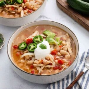 A bowl of chicken chili with white beans, diced tomatoes, and shredded chicken, topped with jalapeรฑos, avocado, cheese, sour cream, and cilantroโperfect for Meal Plan 94. Served with a striped napkin and spoon beside the bowl.