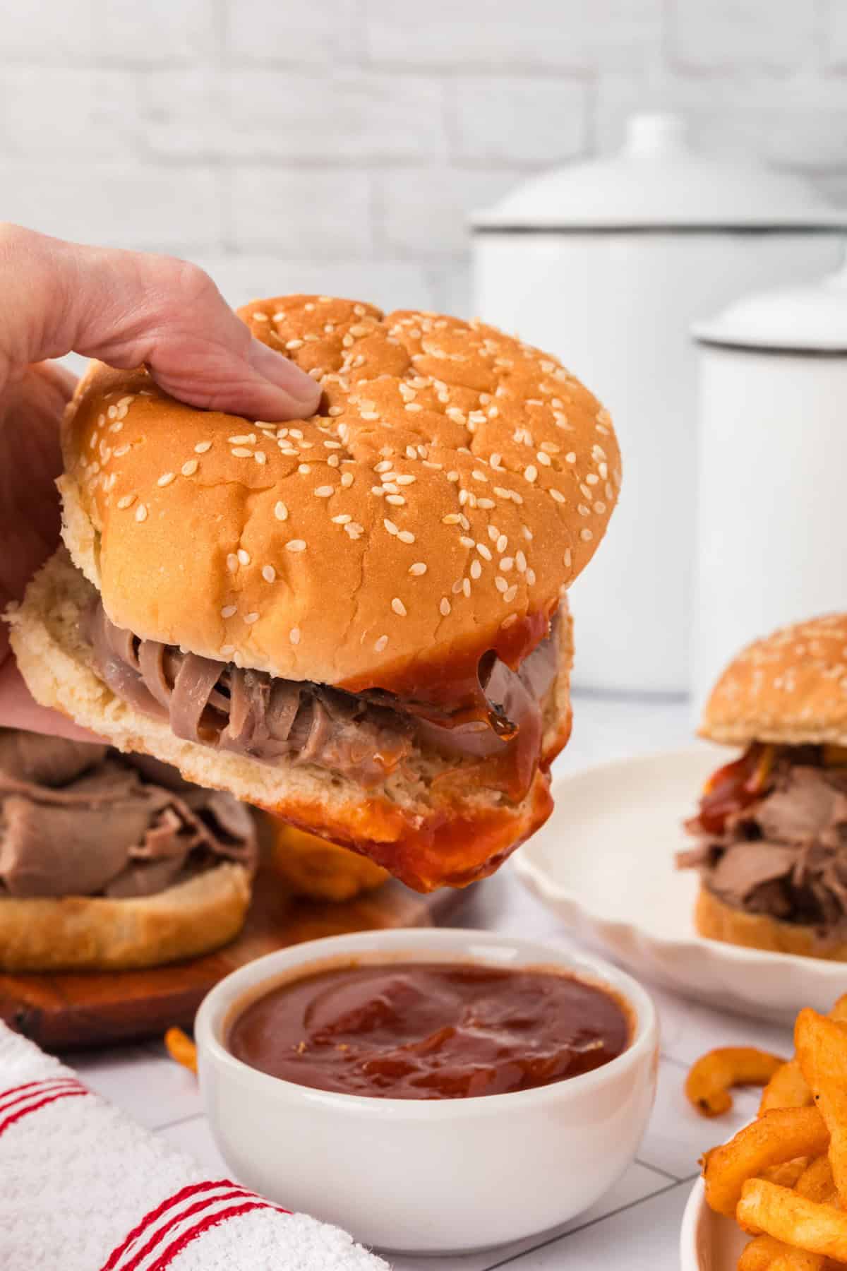 A hand holds a roast beef sandwich with Arby’s Sauce in a sesame bun. Curly fries and a bowl of extra sauce are in the foreground, with another sandwich and kitchen containers in the background.