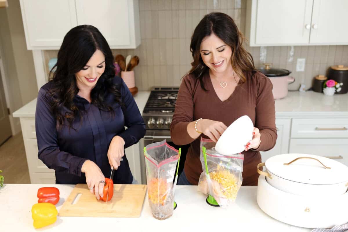 Carrie and Christina prepare food in a bright kitchen; one slices a red bell pepper on a cutting board, while the other adds cheese to a plastic bag with other ingredients. Various kitchen items are around them.