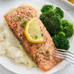 A plate with Lemon Garlic Salmon topped with lemon slices and herbs, served on mashed potatoes with a side of steamed broccoli. A fork rests on the plate.