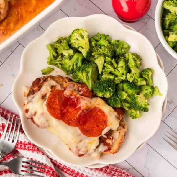 A white plate with a serving of broccoli and a piece of chicken covered in melted cheese and pepperoni slices—perfect for your Meal Plan 90. The plate sits on a tiled surface near a red dish towel and some forks.