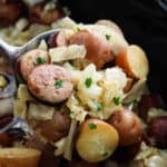 A close-up of Slow Cooker Cabbage and Sausage features a spoonful of cooked sliced sausage, red potatoes, and chopped cabbage, all garnished with parsley. The background reveals more of this comforting dish.