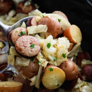 A close-up of Slow Cooker Cabbage and Sausage features a spoonful of cooked sliced sausage, red potatoes, and chopped cabbage, all garnished with parsley. The background reveals more of this comforting dish.