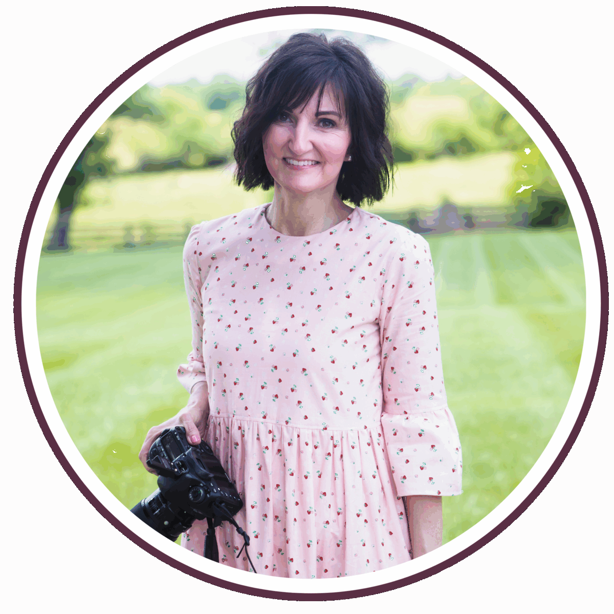 Carrie, with short dark hair, wears a light pink dress with small floral prints as she stands outdoors on green grass holding a camera, smiling at the camera. Trees and a fence are visible in the blurred background.