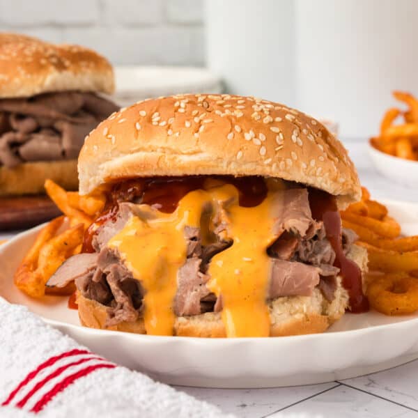 An Arby's Beef and Cheddar sandwich with sliced roast beef, melted cheese, and barbecue sauce on a sesame seed bun is served on a plate with curly fries. Another sandwich and fries appear in the background.
