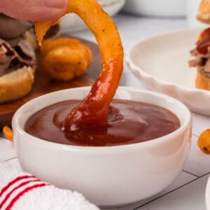 A hand dips a curly seasoned French fry into a small white bowl filled with Arby’s Sauce. A sandwich and more curly fries are visible in the background.