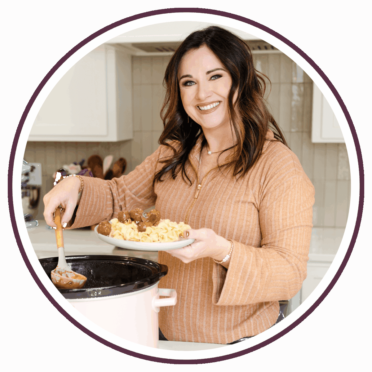 Carrie, a smiling woman with brown hair, serves food from a slow cooker into a bowl while standing in a kitchen. She wears a light brown, long-sleeved top and holds a spoonful of meatballs over pasta.