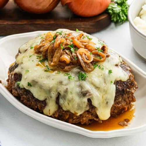 Crockpot French Onion Meatloaf topped with melted cheese and caramelized onions, garnished with chopped parsley, sits on a white platter. A side of mashed potatoes and whole onions are visible in the background.