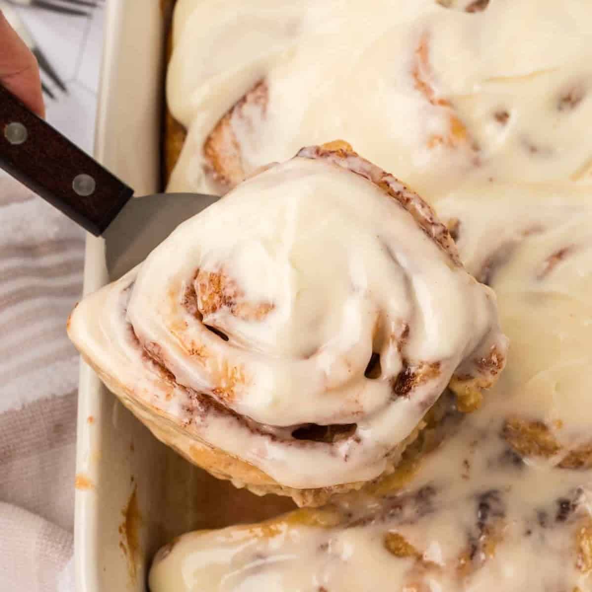 A close-up of Tiktok Cinnamon Rolls covered in creamy white icing being lifted from a baking dish filled with more iced cinnamon rolls.
