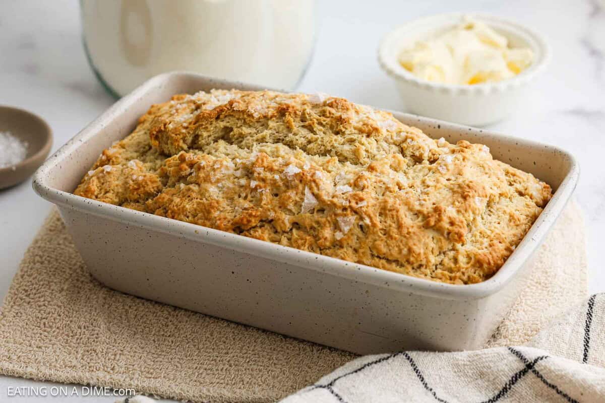 A freshly baked loaf of bread with a golden, textured crust sits in a rectangular baking pan on a beige cloth, with a bowl of butter and a jug of milk in the background.