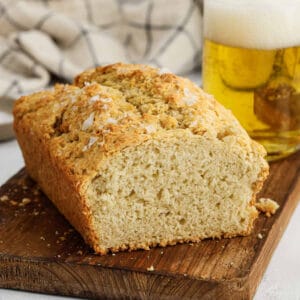 A loaf of homemade bread with a golden crust sits on a wooden board, partially sliced. A glass of frothy beer and a checkered cloth are in the background.