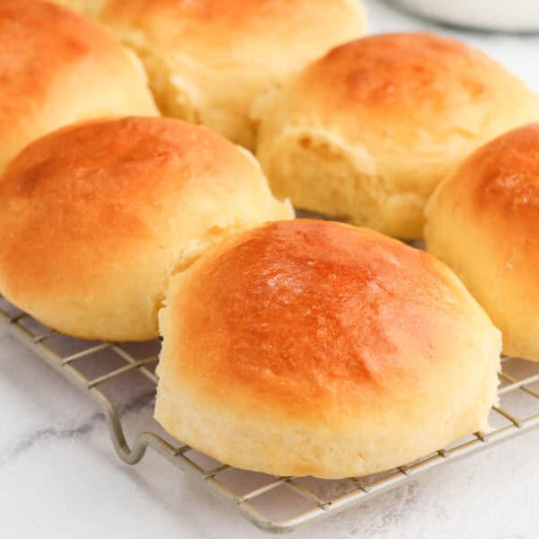 A close-up of six golden brown, freshly baked bread rolls cooling on a metal wire rack with a light background.