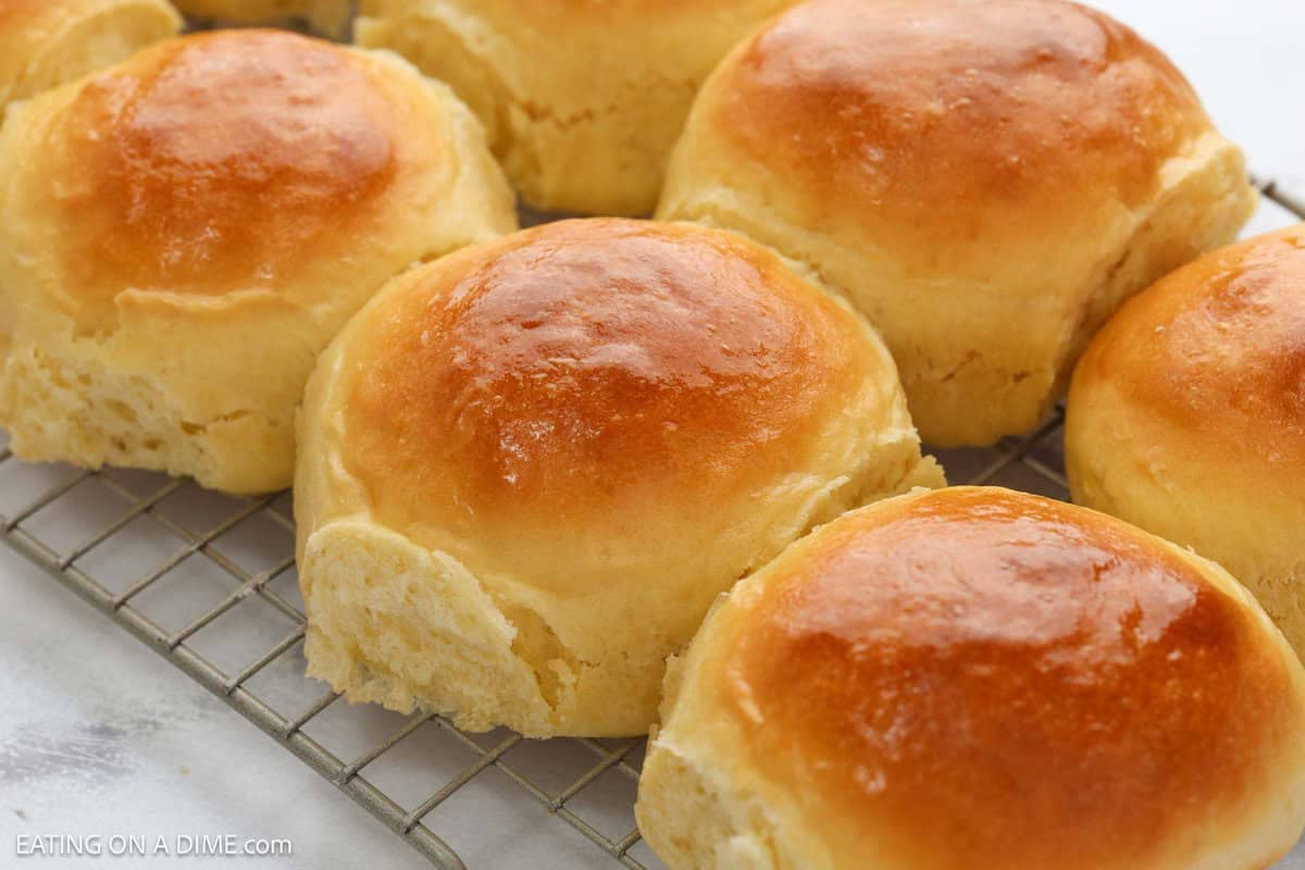 A close-up of several golden brown, freshly baked dinner rolls cooling on a wire rack. The rolls have a shiny, glazed surface and look soft and fluffy.