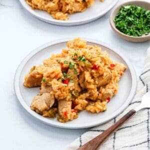 A plate of creamy chicken and rice with chopped vegetables, garnished with fresh herbs. Part of Meal Plan 98, it's served with a bowl of chopped parsley and another plate nearby, alongside a fork and a striped napkin.