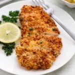 Sliced, crispy breaded chicken cutlets on a white plate, garnished with fresh parsley and a lemon slice, with a fork on the side.