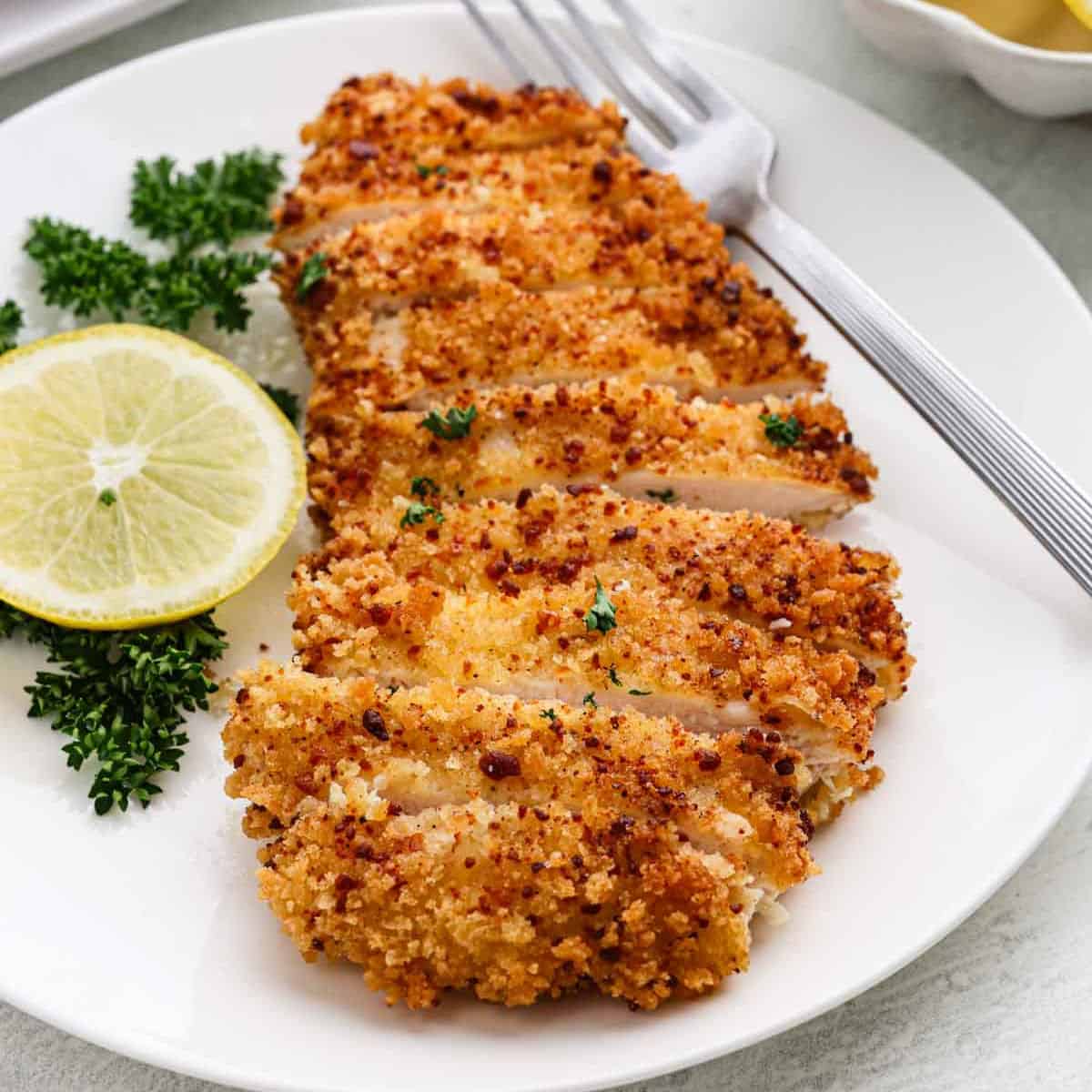 Sliced, crispy breaded chicken cutlets on a white plate, garnished with fresh parsley and a lemon slice, with a fork on the side.