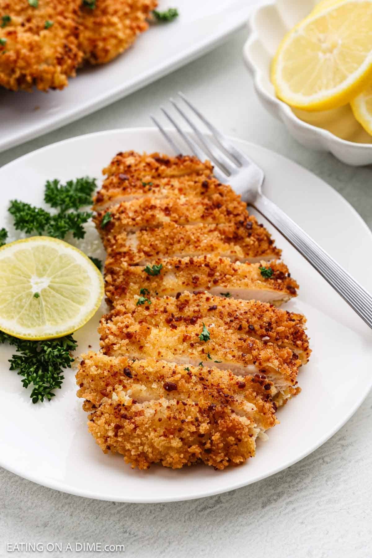 A plate with a sliced, golden-brown breaded chicken cutlet, garnished with a lemon slice and parsley, with a fork beside it. A small bowl of lemon slices is in the background.