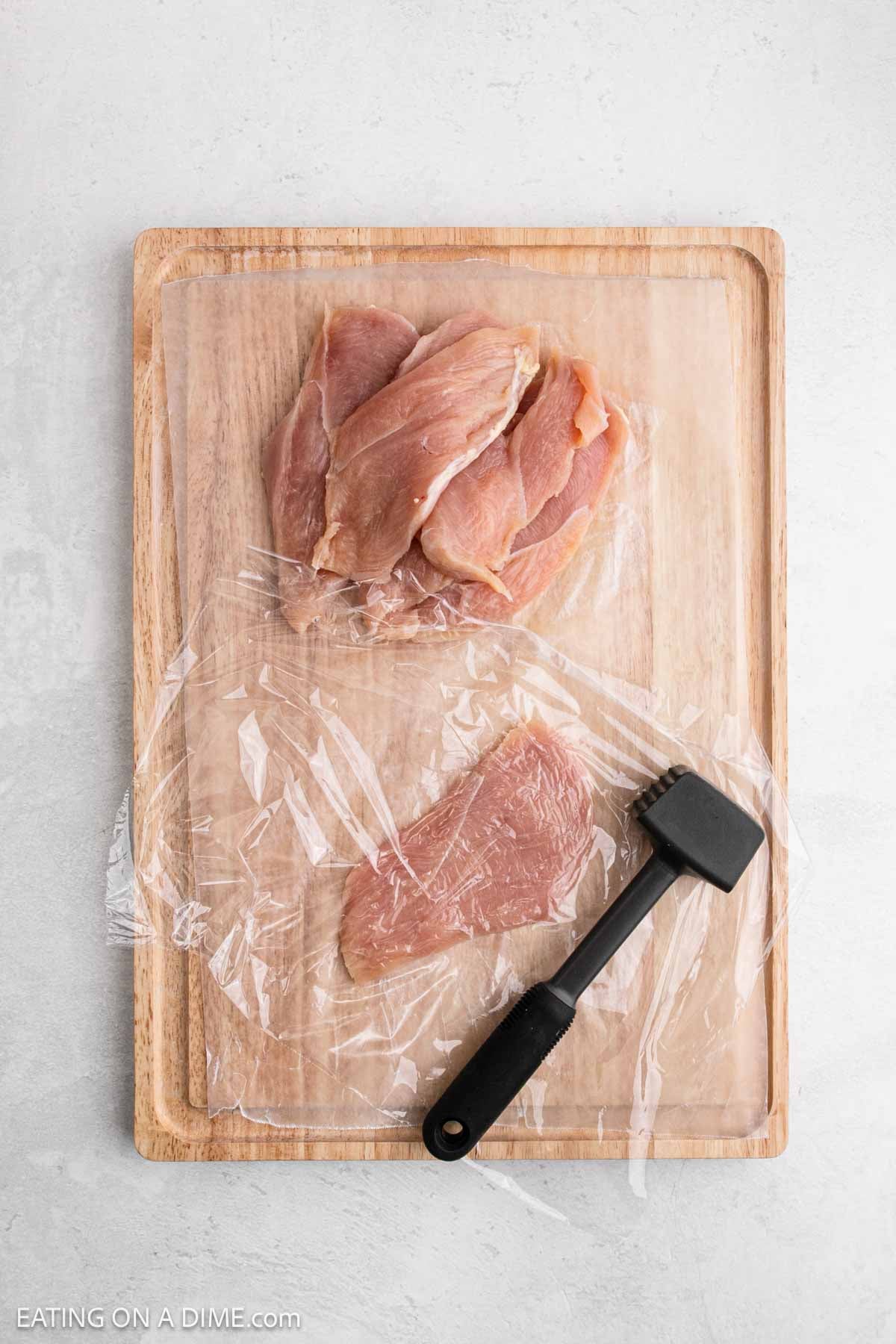 Raw chicken breasts on a wooden cutting board, partially covered with plastic wrap. A meat tenderizer mallet sits beside a chicken breast, ready for pounding into perfect Chicken Cutlets.