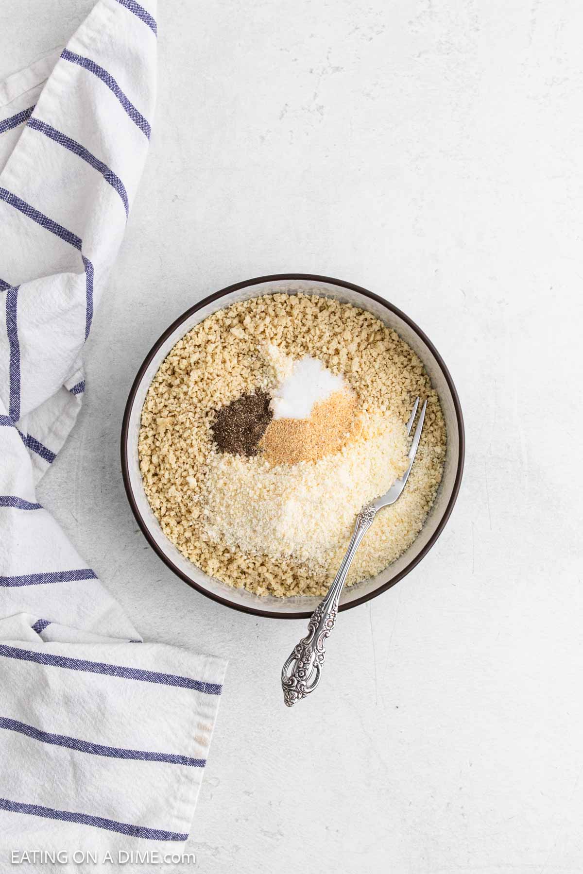 A bowl filled with breadcrumbs and spices for Chicken Cutlets sits on a white surface next to a white and blue striped kitchen towel. A silver fork rests inside the bowl.
