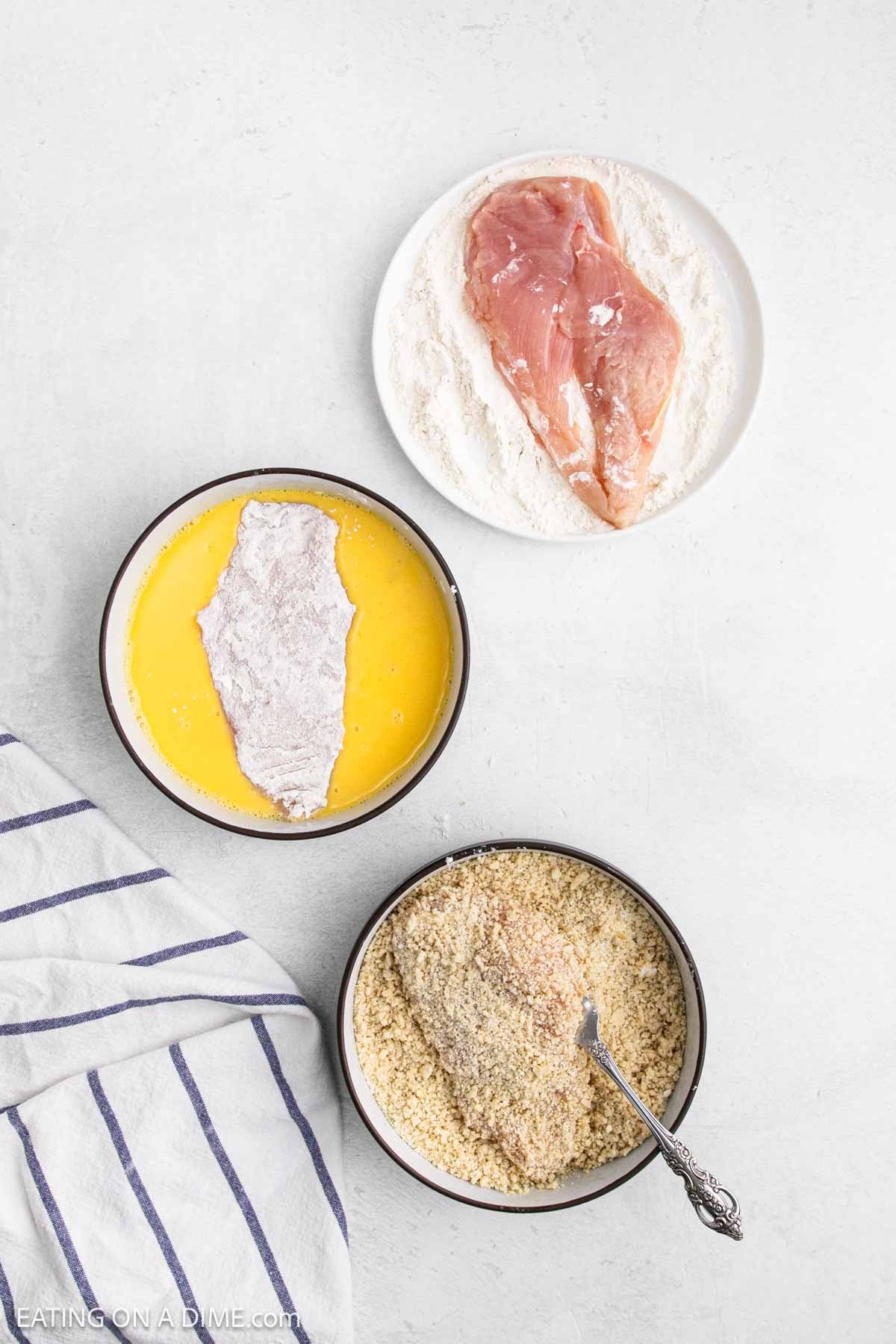 A breading station with raw chicken cutlets on flour, a flour-coated chicken breast in beaten eggs, and another piece being coated in breadcrumbs for breaded chicken, next to a striped kitchen towel.