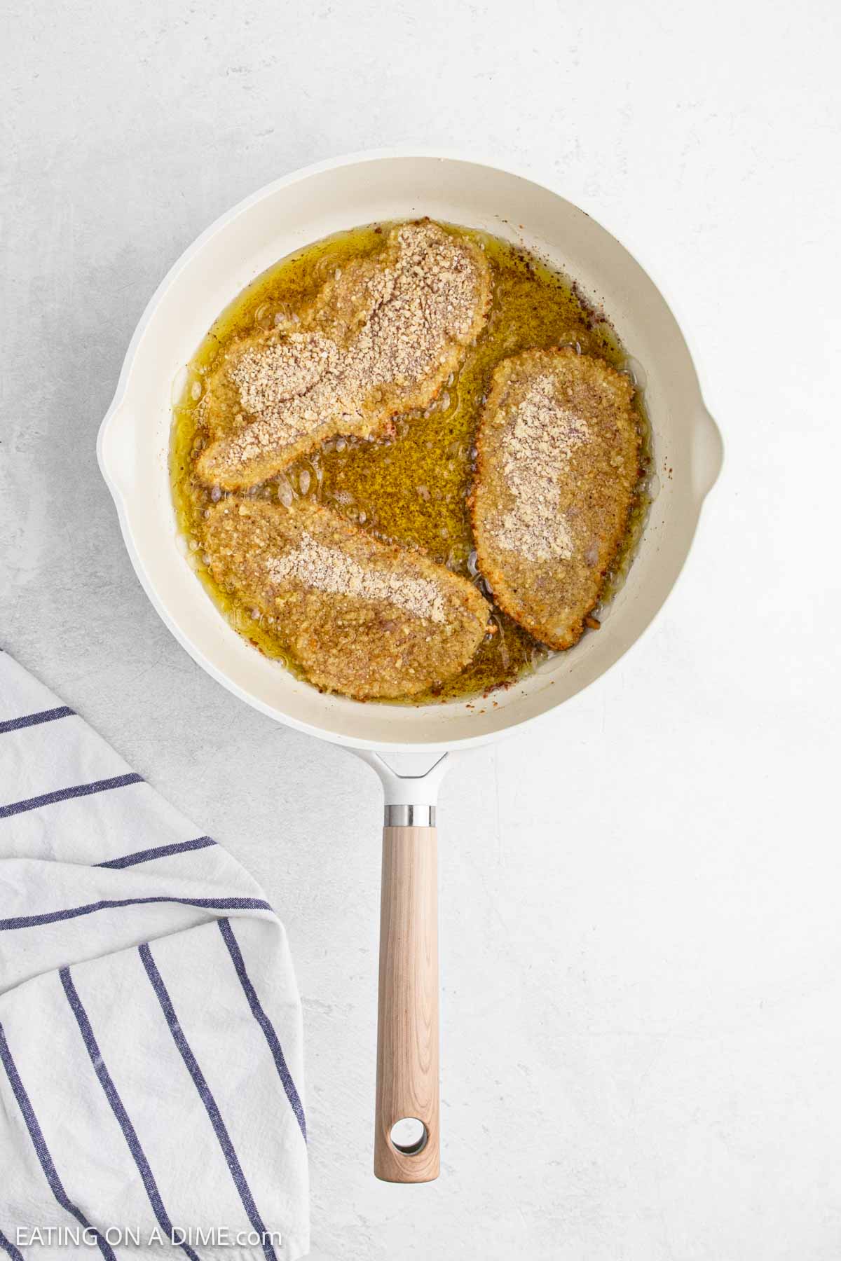 A white skillet with three breaded cutlets frying in oil, perfect for Chicken Recipes, sits next to a white towel with blue stripes on a light surface.