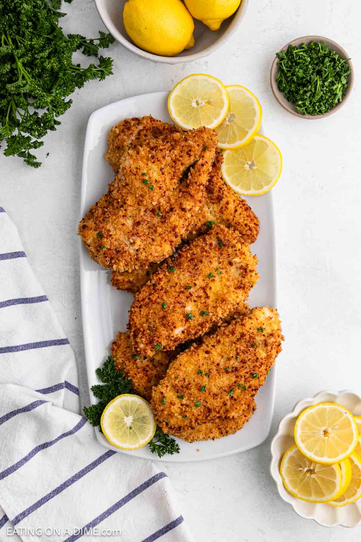 A white platter with crispy breaded chicken cutlets made from tender chicken breast, garnished with parsley and lemon slices, sits on a white surface. Fresh parsley, whole lemons, extra lemon slices, and a striped towel surround the platter.