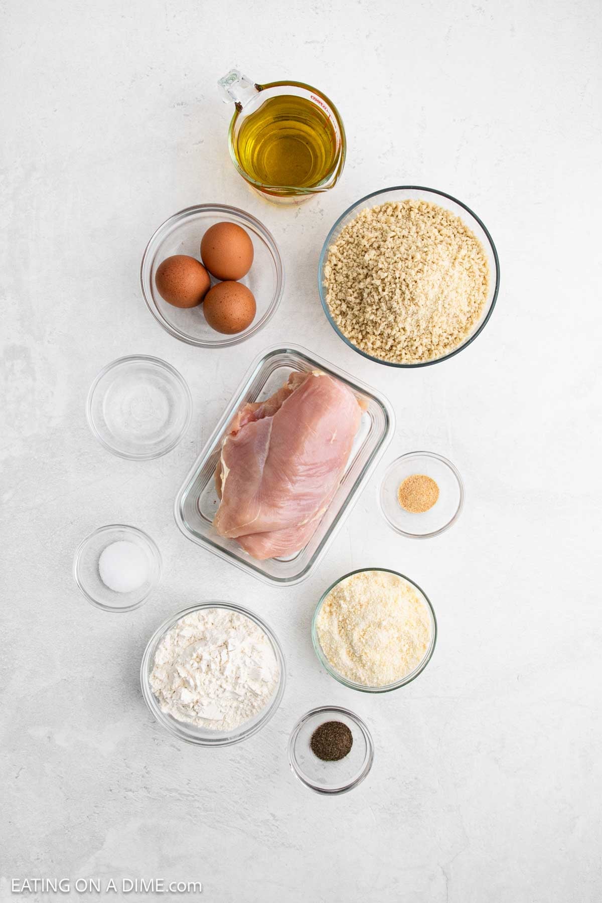 Assorted chicken parmesan ingredients on a white surface, including raw chicken cutlets, eggs, breadcrumbs, olive oil, flour, grated parmesan, salt, pepper, and seasonings in separate glass bowls.