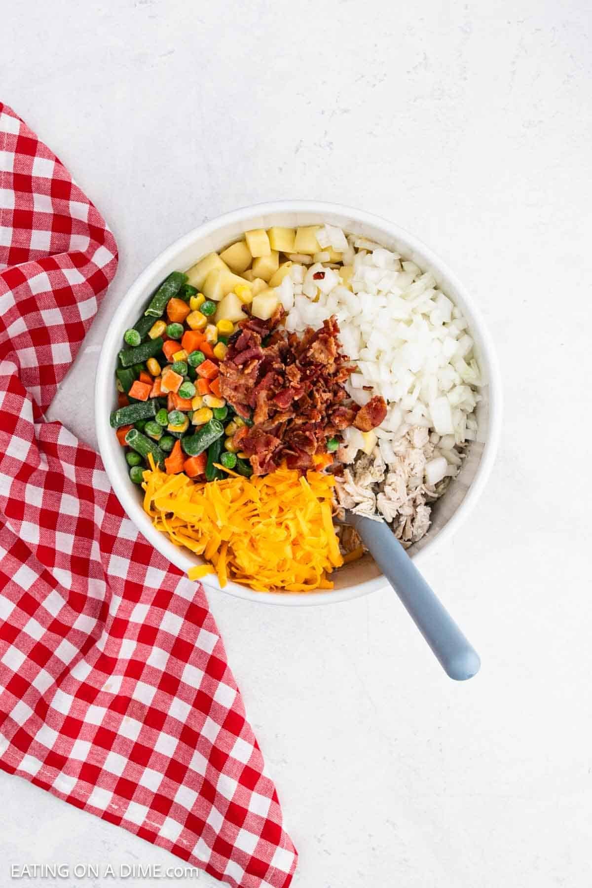 Shredded cheese, mixed vegetables, diced potatoes, chopped bacon, cooked chicken, and diced onions in a bowl with a rubber spoon.