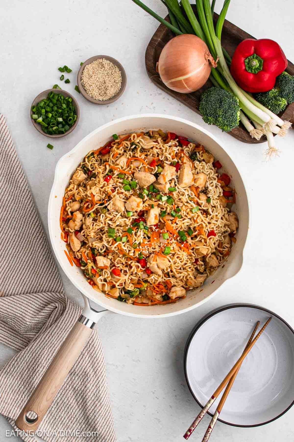 A skillet filled with Chicken Ramen noodles, tender chicken, and vegetables sits on a white surface. Nearby are a bowl of sesame seeds, chopped green onions, broccoli, red bell pepper, a whole onion, and plates with chopsticks.