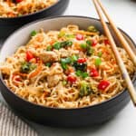 A bowl of Chicken Ramen stir-fry with pieces of chicken, broccoli, red bell peppers, carrots, and green onions, garnished with sesame seeds and served with chopsticks.