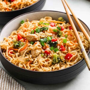 A bowl of Chicken Ramen stir-fry with pieces of chicken, broccoli, red bell peppers, carrots, and green onions, garnished with sesame seeds and served with chopsticks.