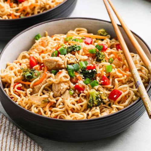 A bowl of Chicken Ramen stir-fry with pieces of chicken, broccoli, red bell peppers, carrots, and green onions, garnished with sesame seeds and served with chopsticks.