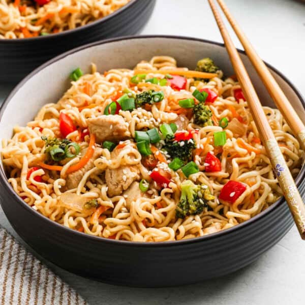 A bowl of Chicken Ramen stir-fry with pieces of chicken, broccoli, red bell peppers, carrots, and green onions, garnished with sesame seeds and served with chopsticks.