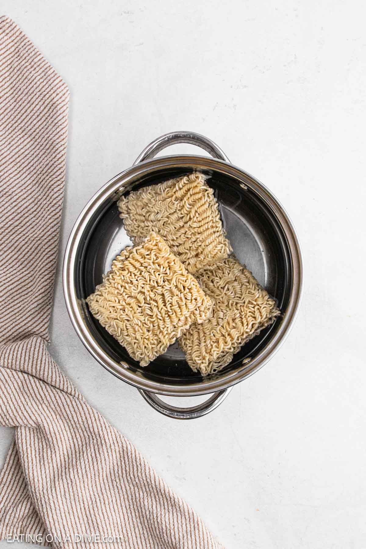 A top-down view of three blocks of uncooked Chicken Ramen noodles placed in a stainless steel pot, with a striped beige towel partially visible beside the pot on a white surface.