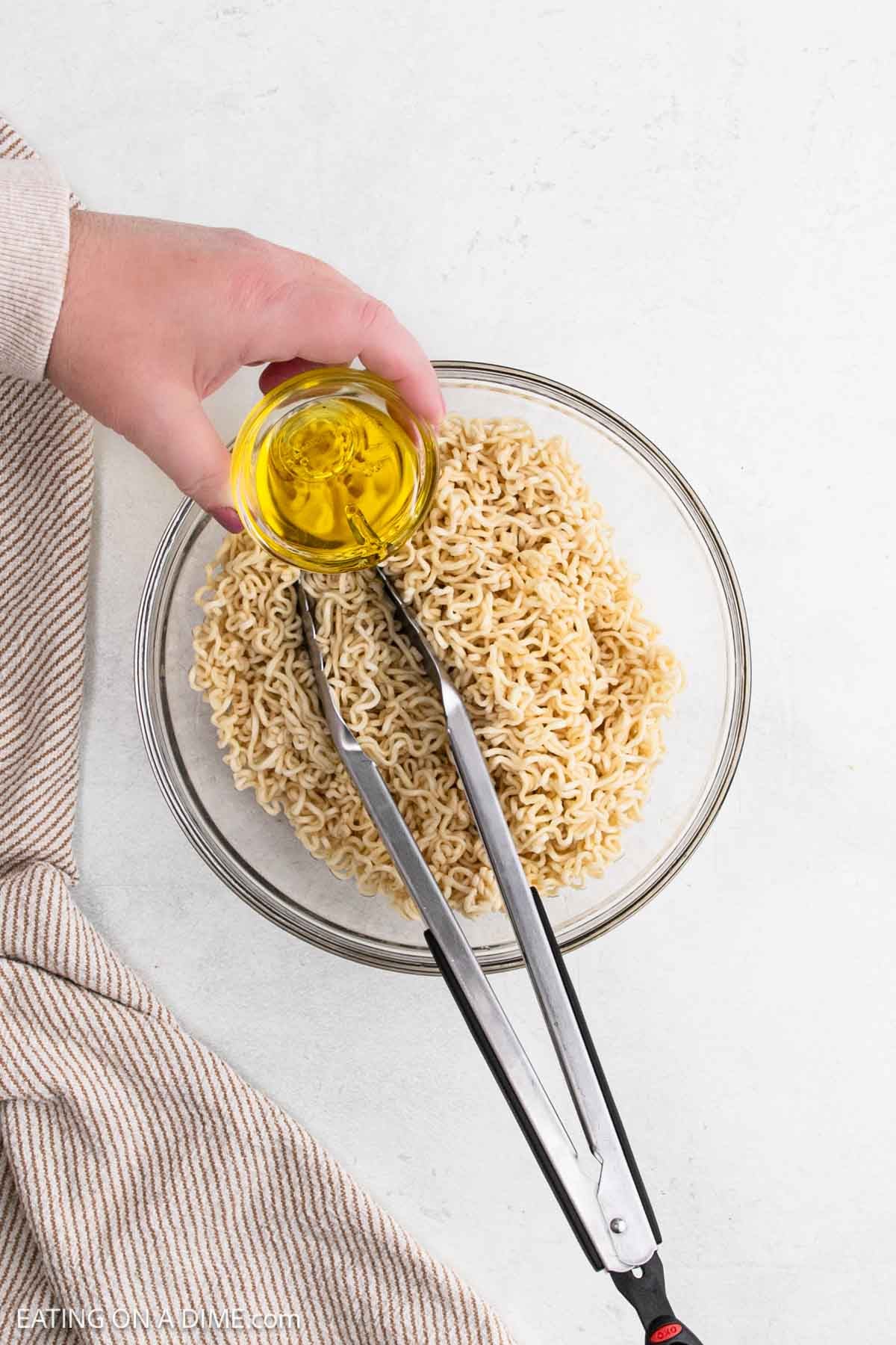 A hand pours olive oil from a small glass bowl over cooked chicken ramen noodles in a large glass bowl, with metal tongs resting on the noodles and a striped cloth nearby.